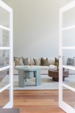 A view through open double glass doors to a living room with comfortable seating, styled with a range of cushions and decor on tables