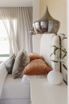 modern, light bedroom with white bedding and accents of neutrals and orange. styled with unique lighting and a plant on the sidetable